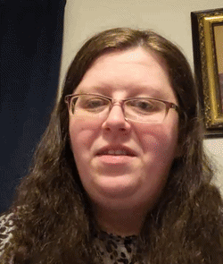 A woman with long wavy brown hair and glasses speaking to the camera.