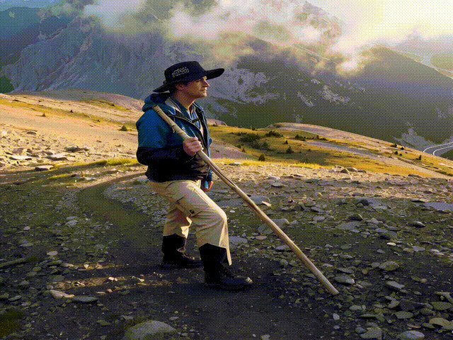 A video of a man standing on a trail above the tree line watching the scenic view.