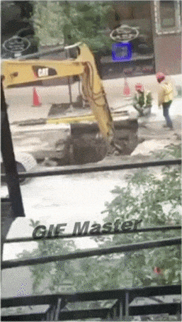 A yellow CAT excavator digging a trench on a city street with a construction worker nearby.