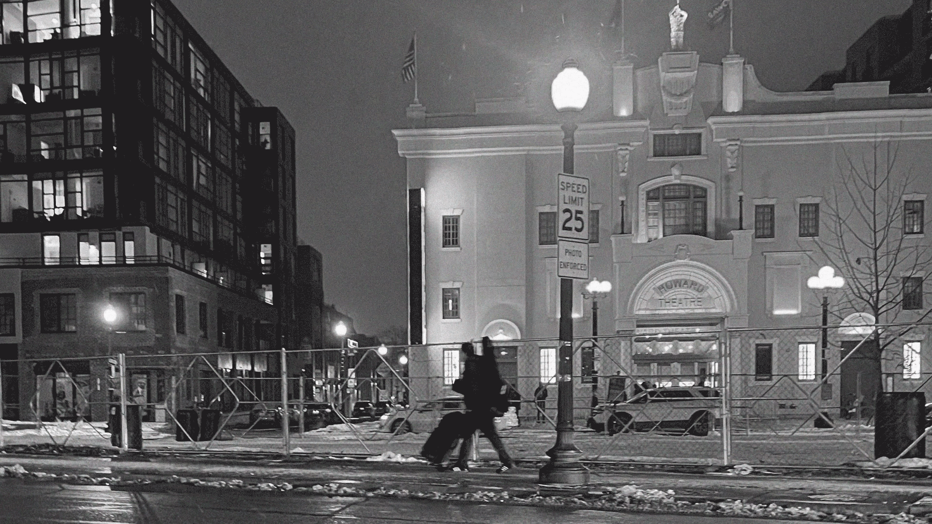 black and white photo of a man walking in a city with a guitar and music gear