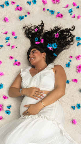 Mujer embarazada sonriente rodeada de flores azules y rosas, celebrando su revelación de género con alegría y ternura.