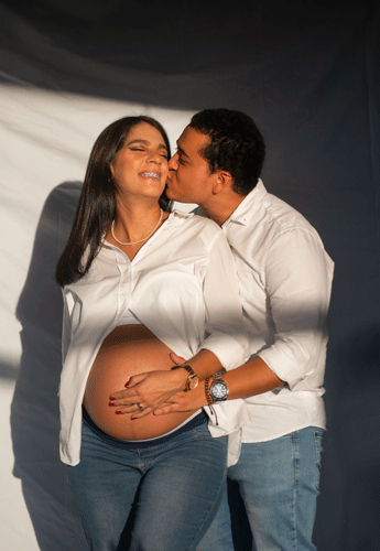 Pareja con mujer embarazada, hombre besando la mejilla de la mujer, mostrando ternura y complicidad.