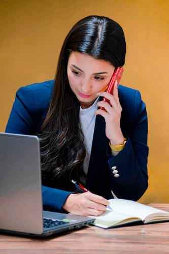 Mujer frente a PC, con agenda en escritorio y celular en el oído, trabajando.
