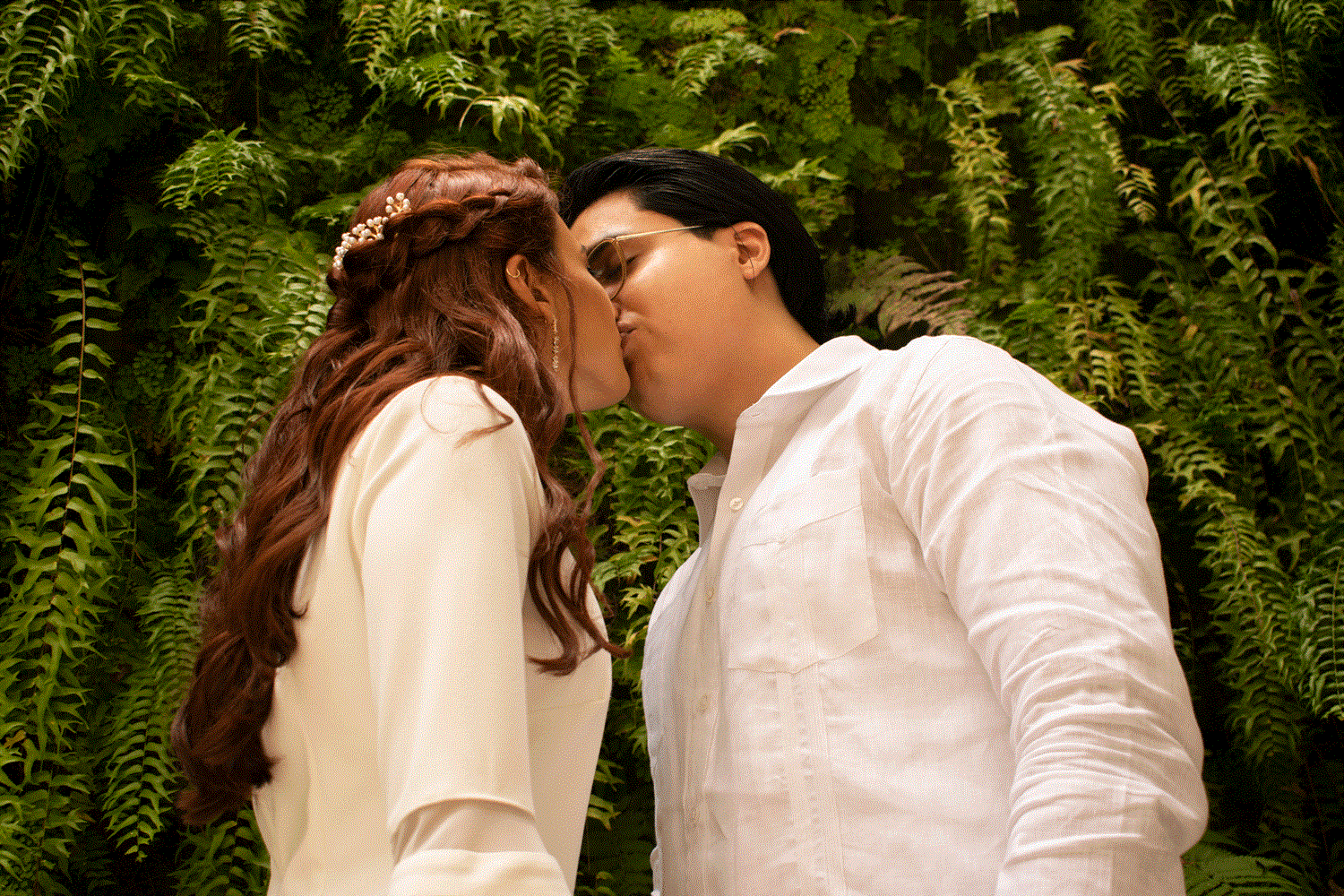 Beso de recién casados sobre fondo de plantas verdes, capturando un momento natural y romántico.