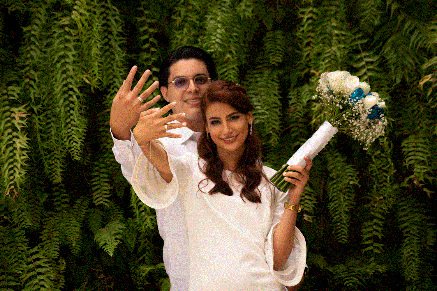 Pareja de recién casados mostrando sus anillos de compromiso, sonrientes, con fondo de plantas verdes.