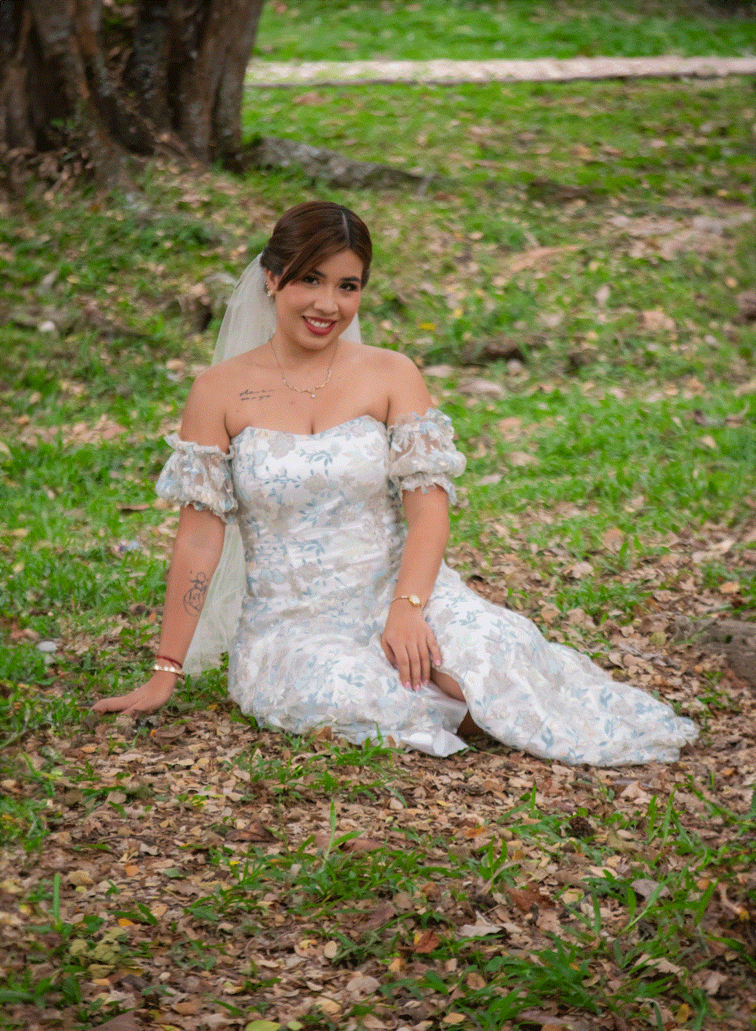 Novia sentada en el pasto, sonriendo con felicidad, capturando un momento relajado y natural en su día especial.