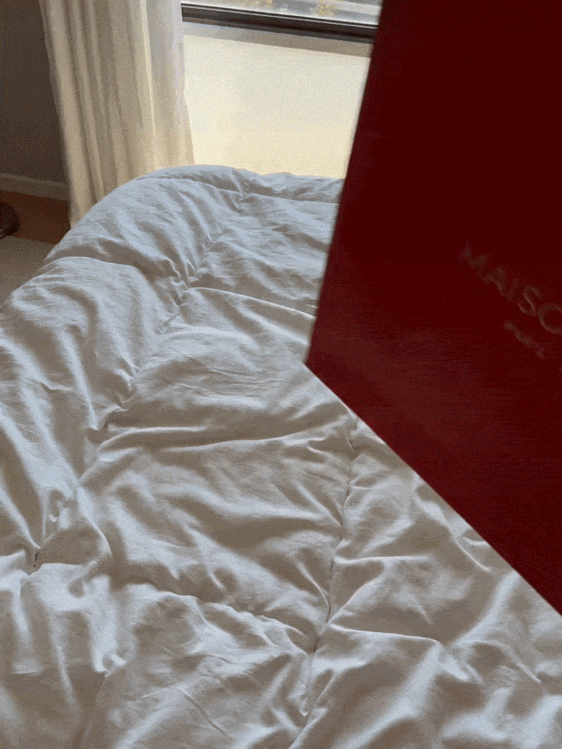 Red Maison Francis Kurkdjian luxury shopping bag on a white quilted duvet bed.