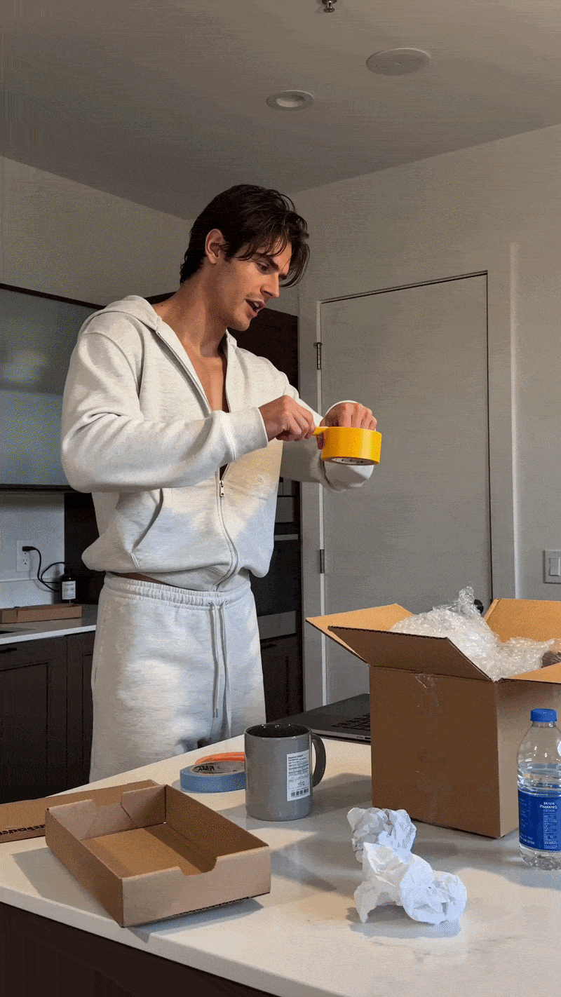 A man in a white sweatsuit uses yellow packing tape to seal a cardboard shipping box on a kitchen island.
