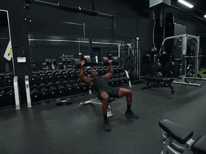 A man performs an incline dumbbell press for chest strength training in a modern gym setting.