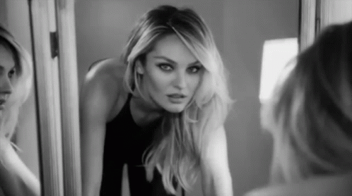 Black and white fashion portrait of a blonde model with smoky eye makeup looking into a mirror.