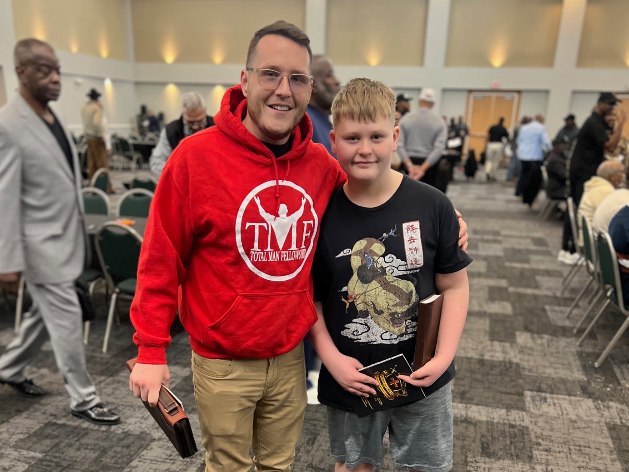 Man in red hoodie posing with a young boy holding a book