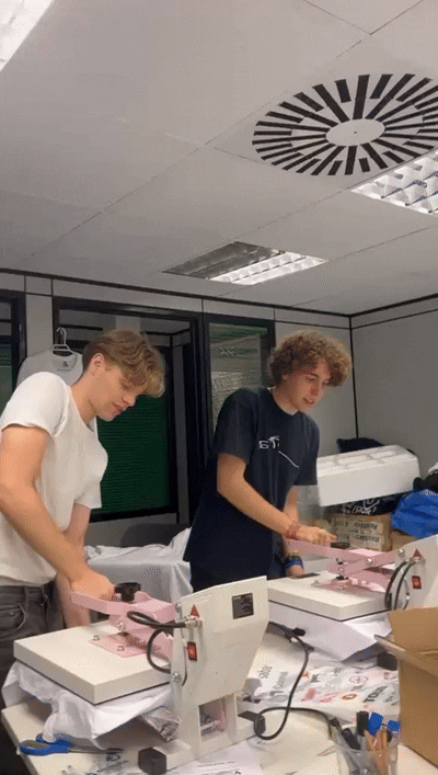 Two young men using professional heat press machines to print custom t-shirt designs in a studio.