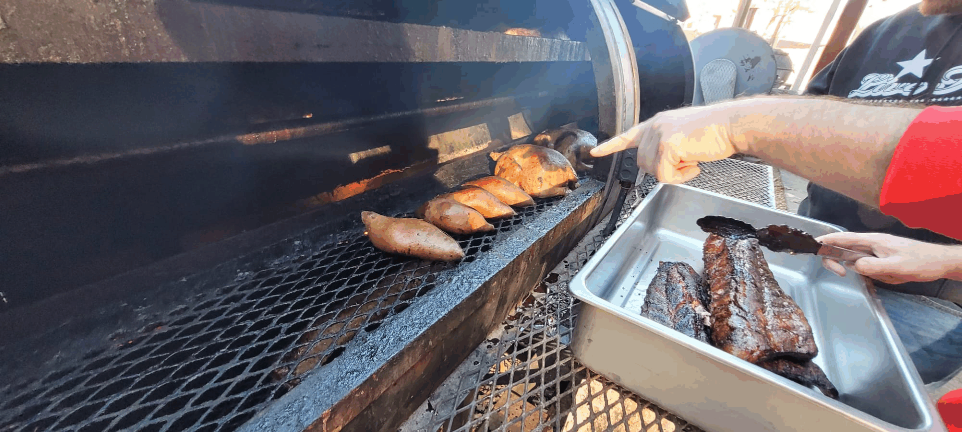 A pitmaster smoking sweet potatoes and pork ribs on a large black outdoor offset smoker grill.