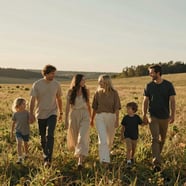 A cinematic, wide-angle lifestyle photograph of a family walking through a sun-drenched meadow in a North American / US countryside setting. The lighting is the warm, golden glow of late afternoon. The subjects are dressed in soft sand and charcoal tones, captured in a candid moment of laughter. The image feels authentic and emotionally resonant.