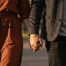 A close-up lifestyle shot of a couple's hands held together during a walk in a North American / US park at golden hour. Warm cinematic lighting with charcoal and terracotta clothing accents.
