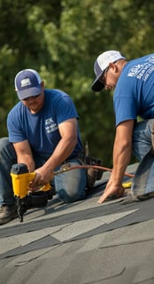 Professional roofing contractors installing asphalt shingles on a residential roof using a pneumatic nail gun.
