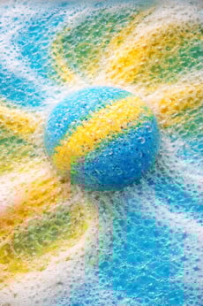 Oversized bath bomb releasing blue and yellow foam in soft, cloud-like fizz.