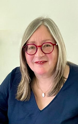 Professional portrait of a smiling woman with blonde hair wearing stylish red glasses and a blue v-neck top.