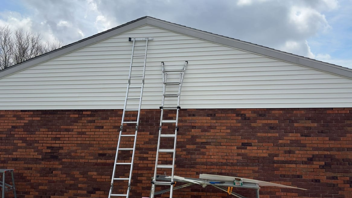 Two aluminum extension ladders leaning against a house with white siding and brick for home repair work.