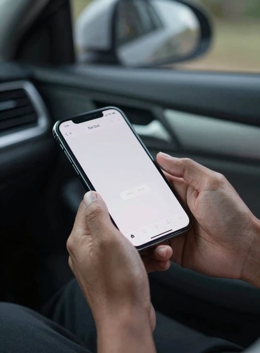 Close-up photography of a Southeast Asian / Indonesian person's hands holding a smartphone with a clean interface, sitting inside a comfortable car, soft evening light, mist grey interior.