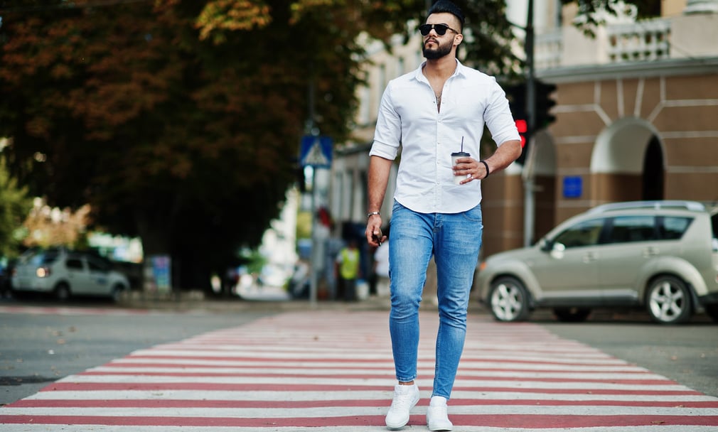 homem atravessa a rua em uma faixa de pedestres. Ele usa uma camisa branca, jeans e tênis branco