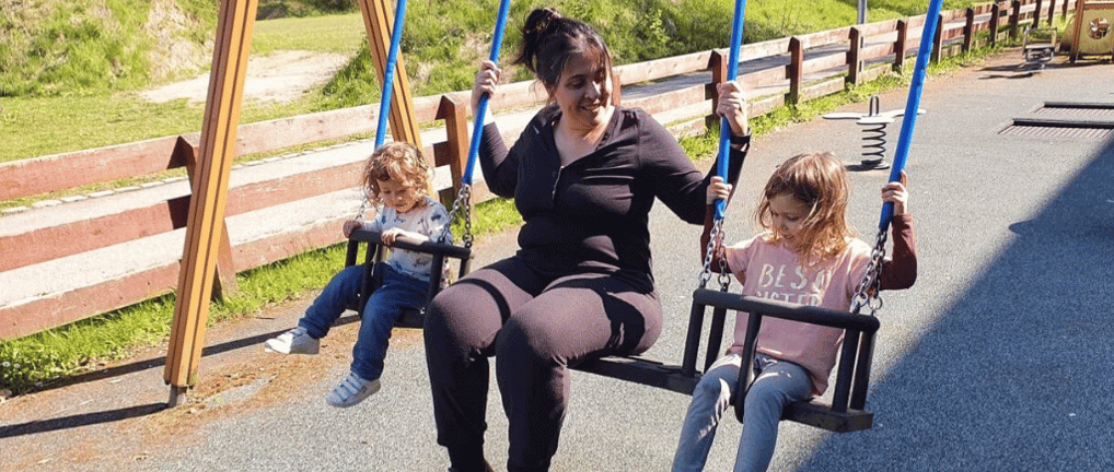 a mom sitting on a swing set with her two bilingual kids