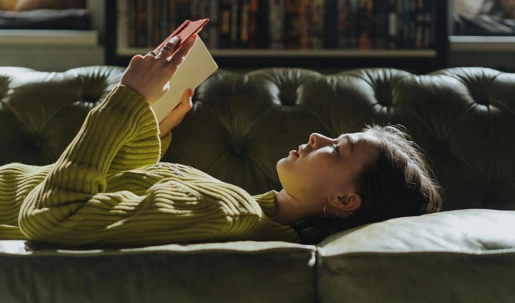 Uma jovem tranquilamente em um sofá, lendo um livro de artes visuais.