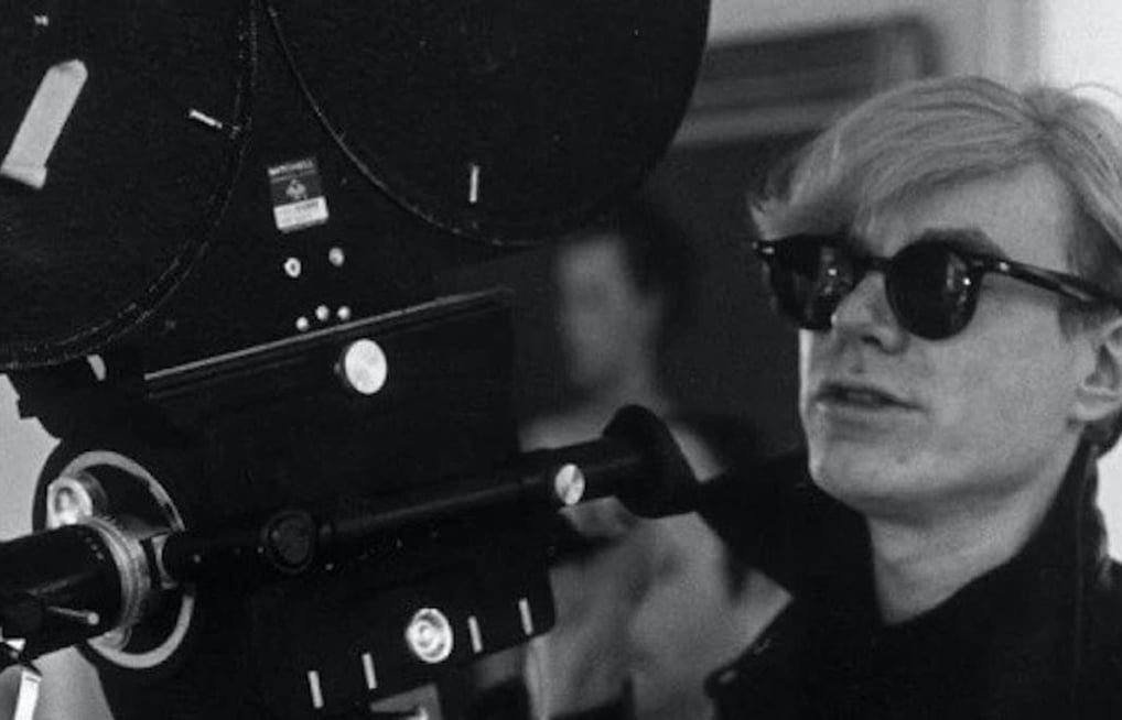 Andy Warhol segurando uma câmera de filme antiga, usando óculos escuros preta