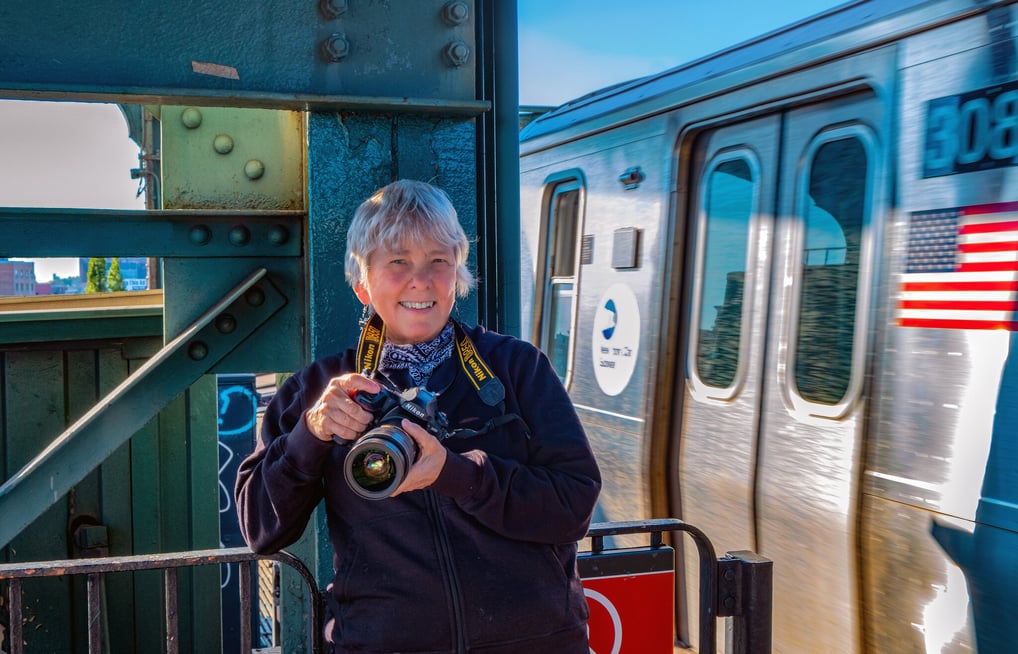 A fotógrafa Martha Cooper, segurando uma câmera e sorrindo, posa em um ambiente urbano