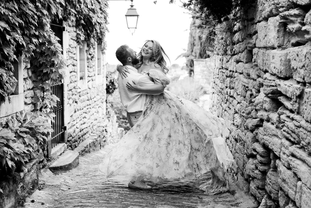 Black and white editorial engagement photo – couple laughing as groom twirls bride in village alley