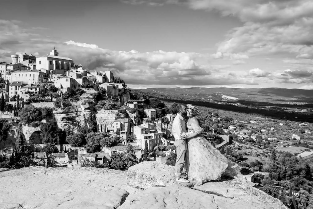 Wide black and white couple photo — kiss on the cheek with panoramic view over Gordes, Provence