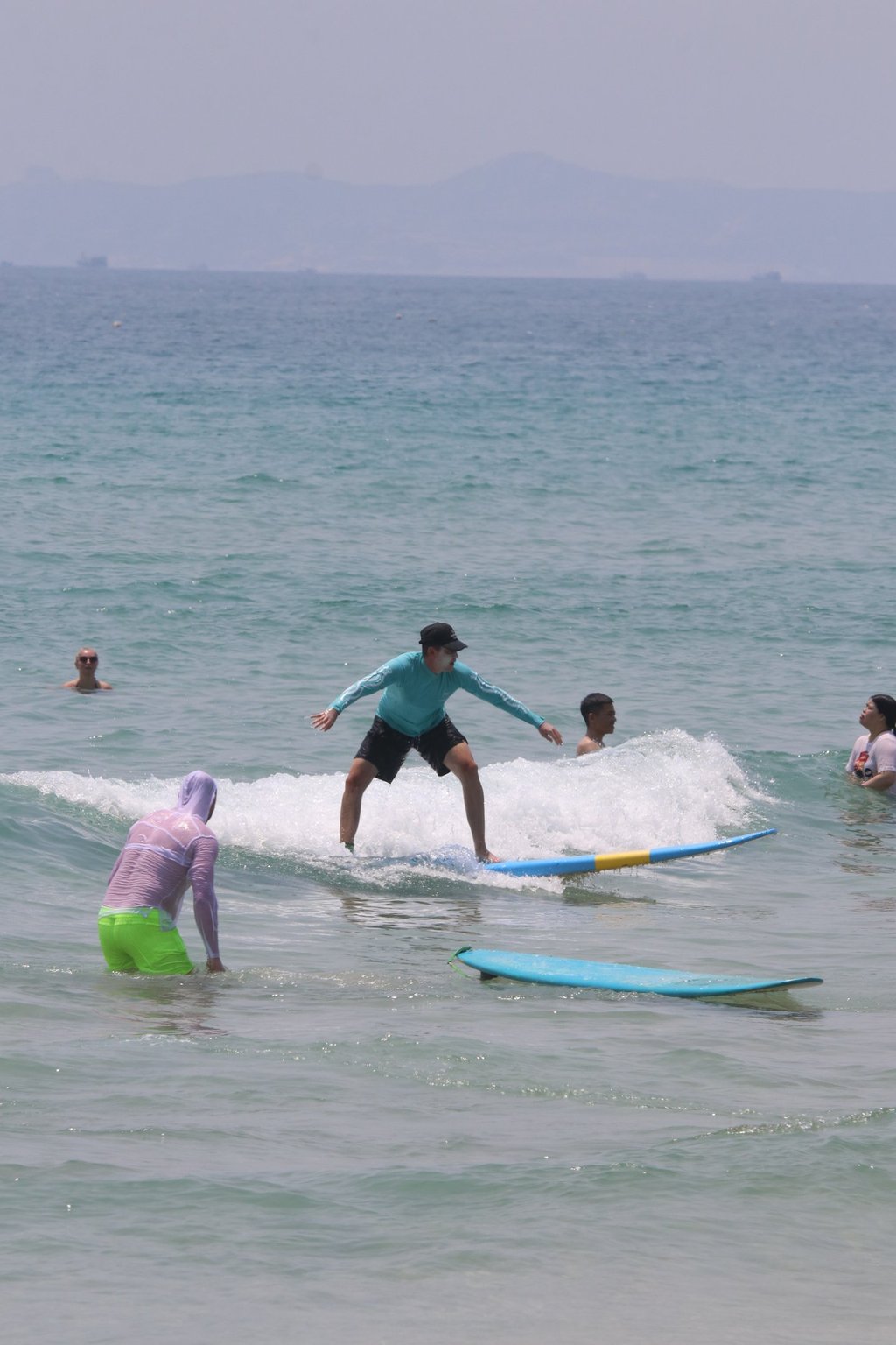 A surfer in a blue rash guard rides a small ocean wave on a surfboard near other swimmers.