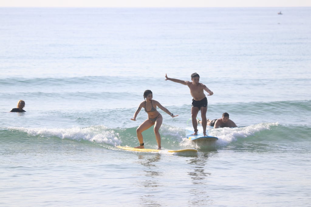 A man and woman riding surfboards on a small ocean wave during a sunny beach surfing lesson.