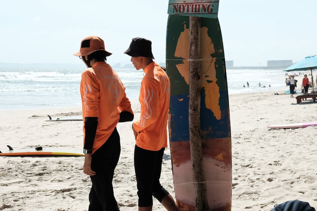 Two surfers in orange shirts and hats stand on a sunny beach next to a surfboard, with ocean waves and people in the