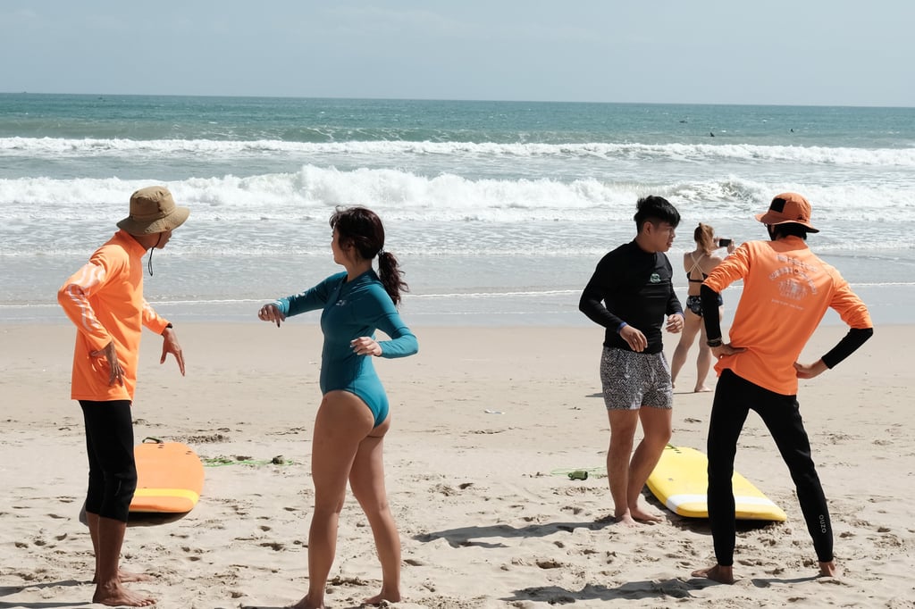 Surfing instructors teaching a group of students on a sunny beach with waves in the background.