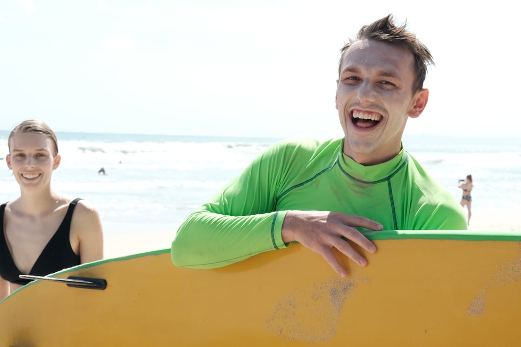 A happy young man in a green rash guard holding a yellow surfboard at a sunny beach.