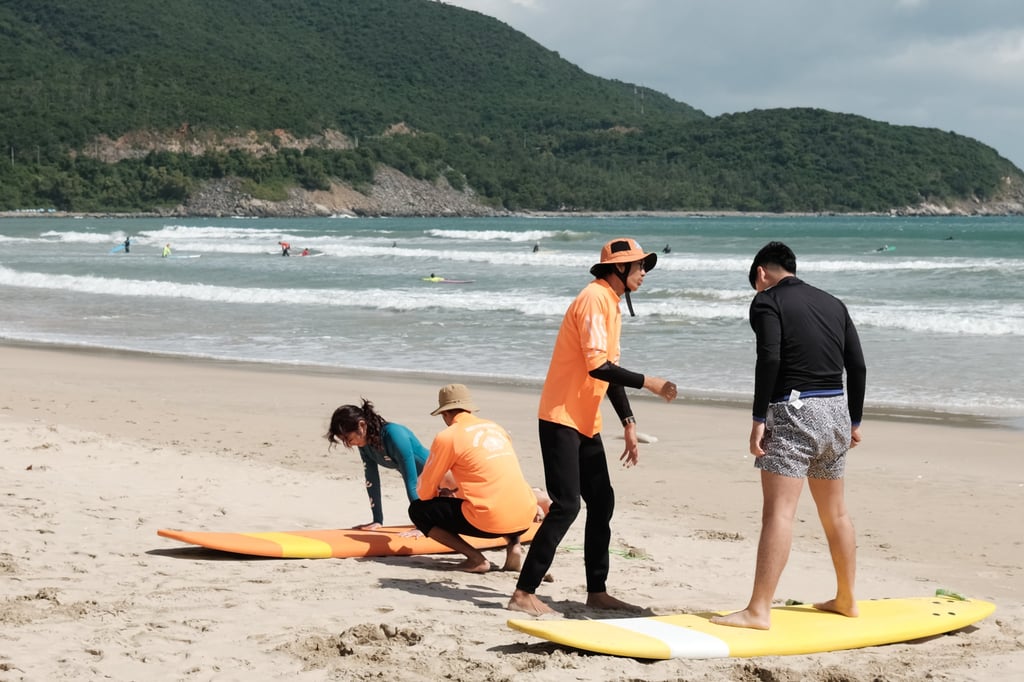 Instructors in orange shirts lead a group surf lesson on a sandy beach with ocean waves.