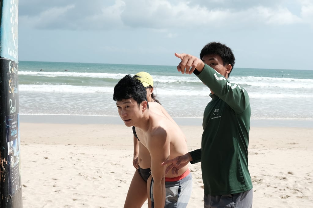 A surf instructor in a green shirt pointing at ocean waves for a student on a sandy beach.