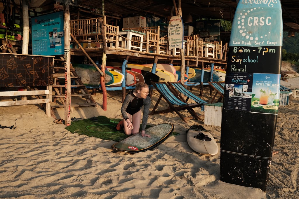 A female surfer preparing her surfboard at Cam Ranh Surf Camp on a sandy beach at sunrise.