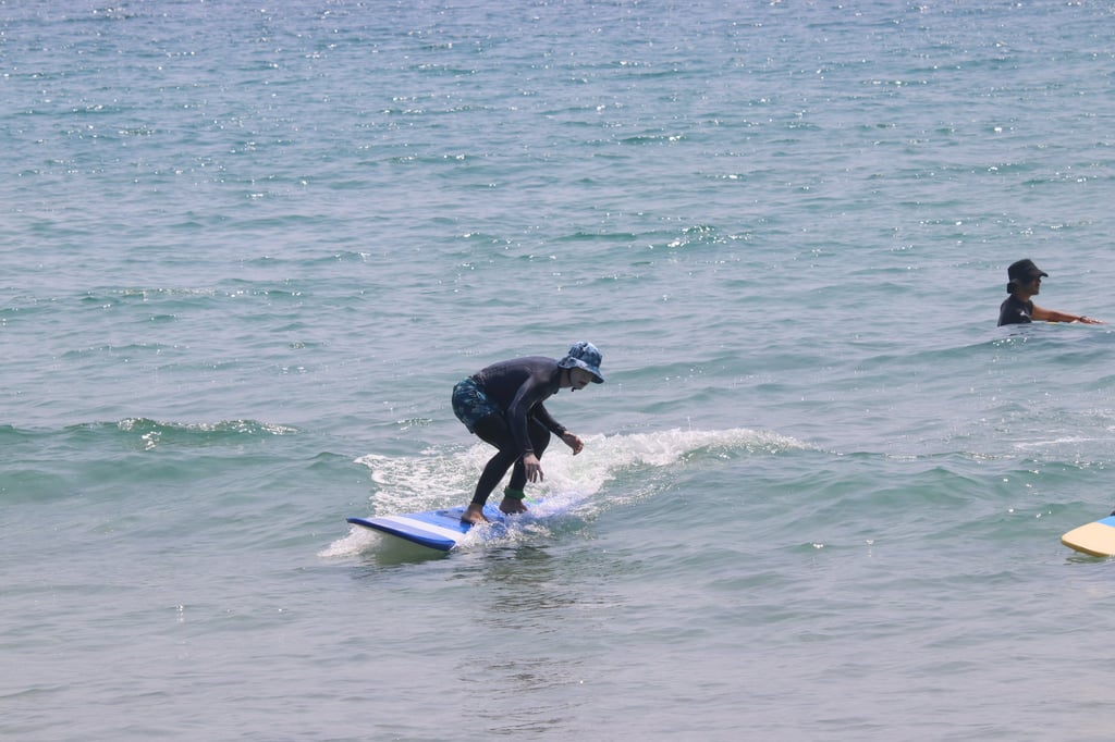 A person in a black wetsuit and blue hat surfs a small wave on a light blue surfboard in the ocean.