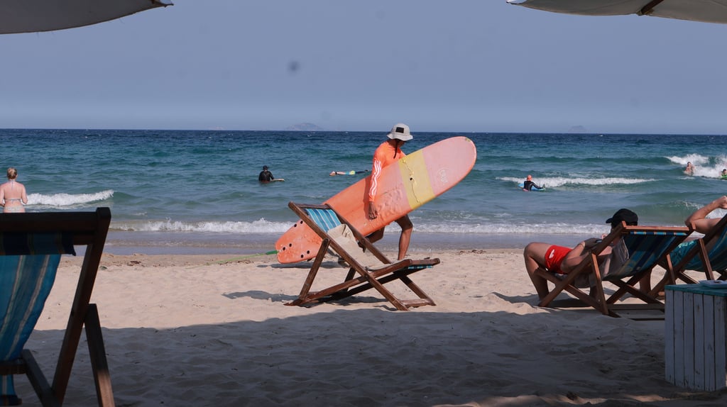 A person in a sun hat carrying a surfboard on a sunny beach with beach chairs and ocean waves.