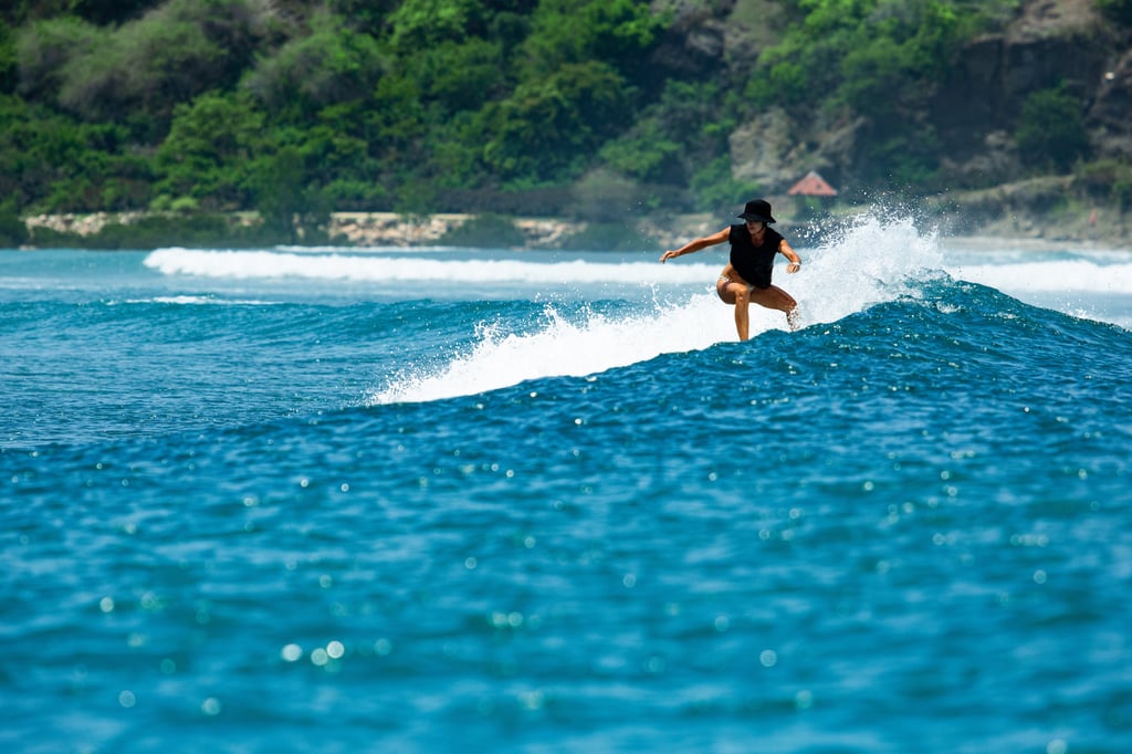 A female surfer in a black swimsuit catches a blue wave near a tropical coastline.
