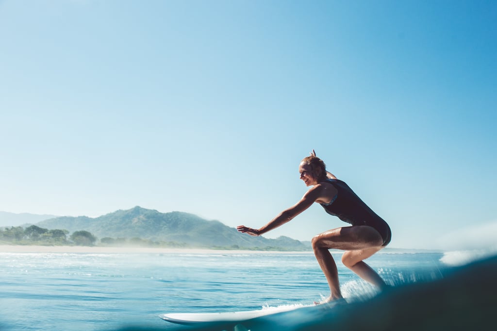A smiling woman in a black swimsuit surfing on a blue ocean wave near a tropical beach coastline.