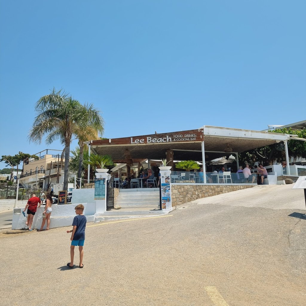 Lee Beach Food, Drinks & Cocktail Bar on a sunny day in Greece with a palm tree and tourists.