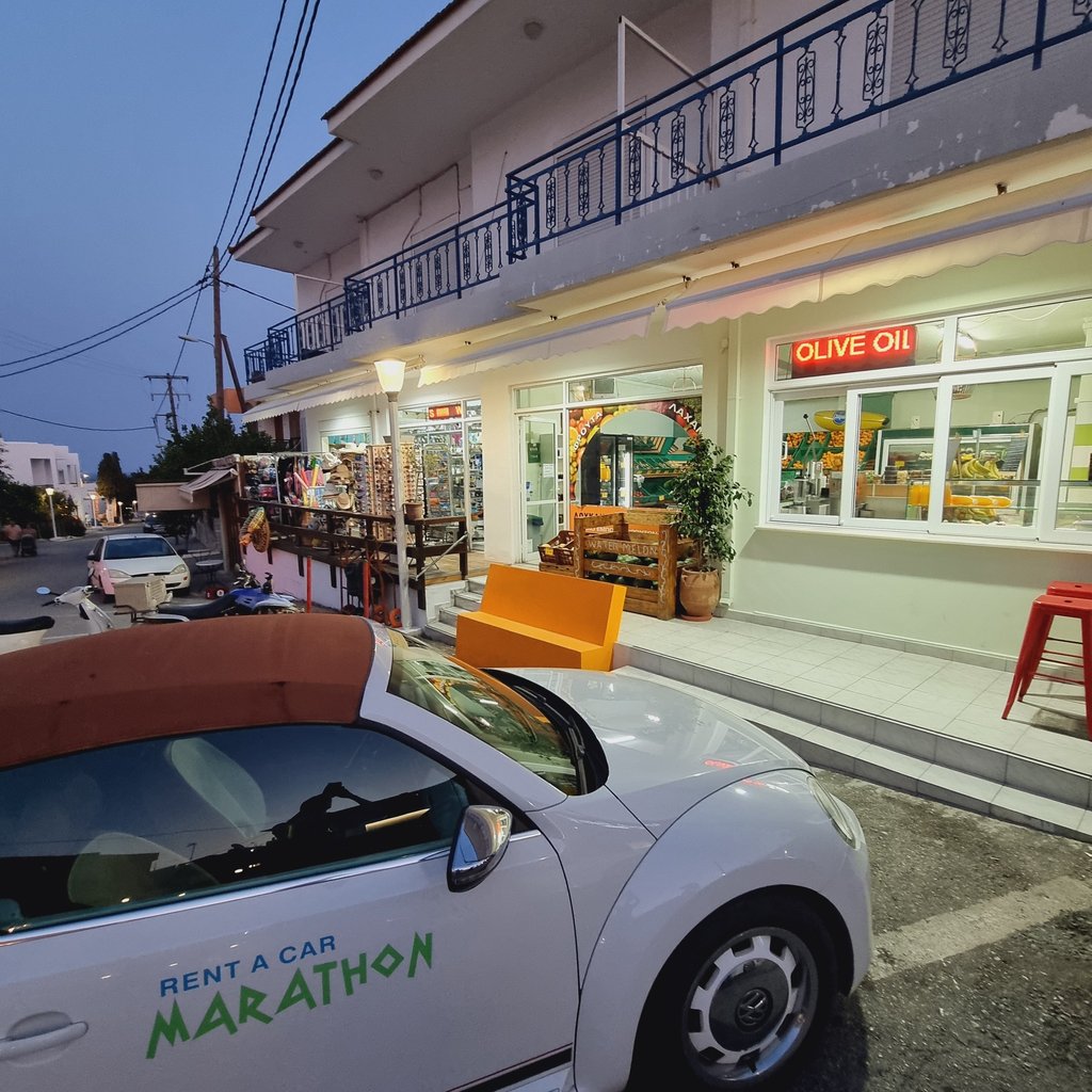 White Marathon rental car parked outside a Greek village market and olive oil shop at twilight.