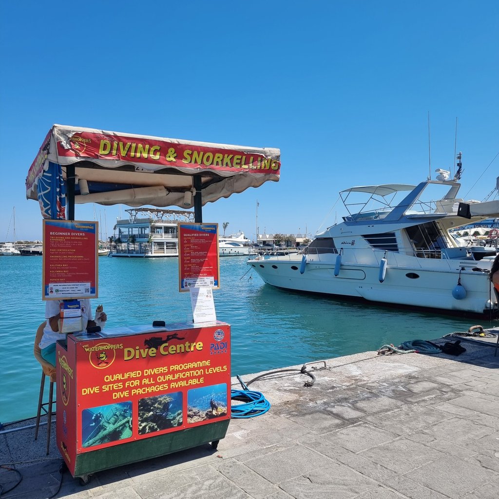 A scuba diving and snorkeling tour kiosk at a sunny Mediterranean harbor with luxury yachts in the background.