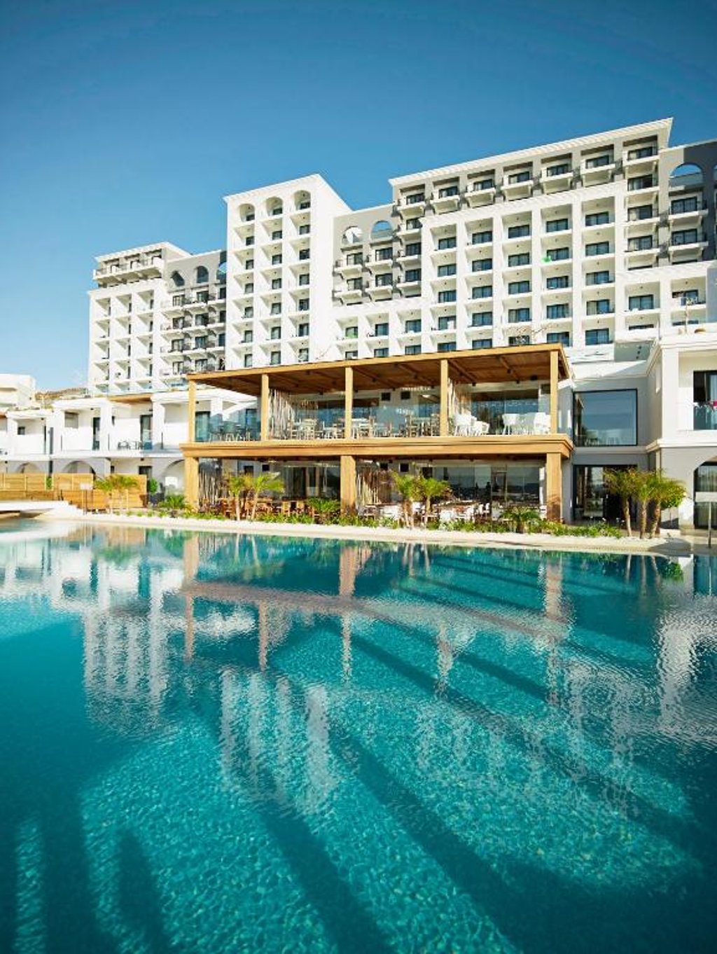 Luxury white resort building with balconies reflecting in a large outdoor swimming pool under a blue sky.