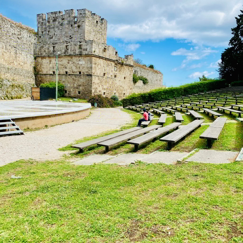 Outdoor stone theater with wooden benches next to the historic Palace of the Grand Master in Rhodes, Greece.