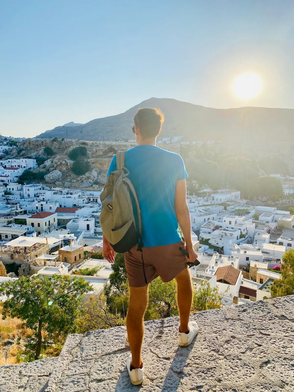 A male traveler with a backpack overlooks the white architecture of Lindos, Rhodes during a sunny Mediterranean sunset.