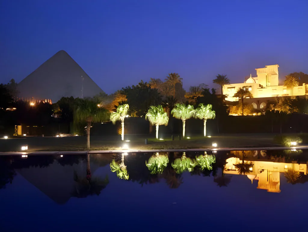 Night view of the Great Pyramid of Giza reflected in a luxury resort pool with palm trees.
